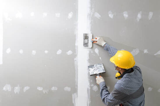 A picture of a man wearing a safety helmet painting a drywall with white paint.