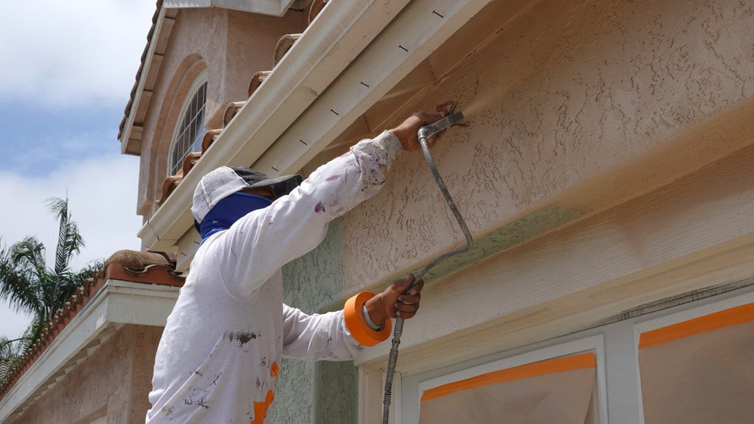 A picture of a man spraying house exterior wall with beige paint.