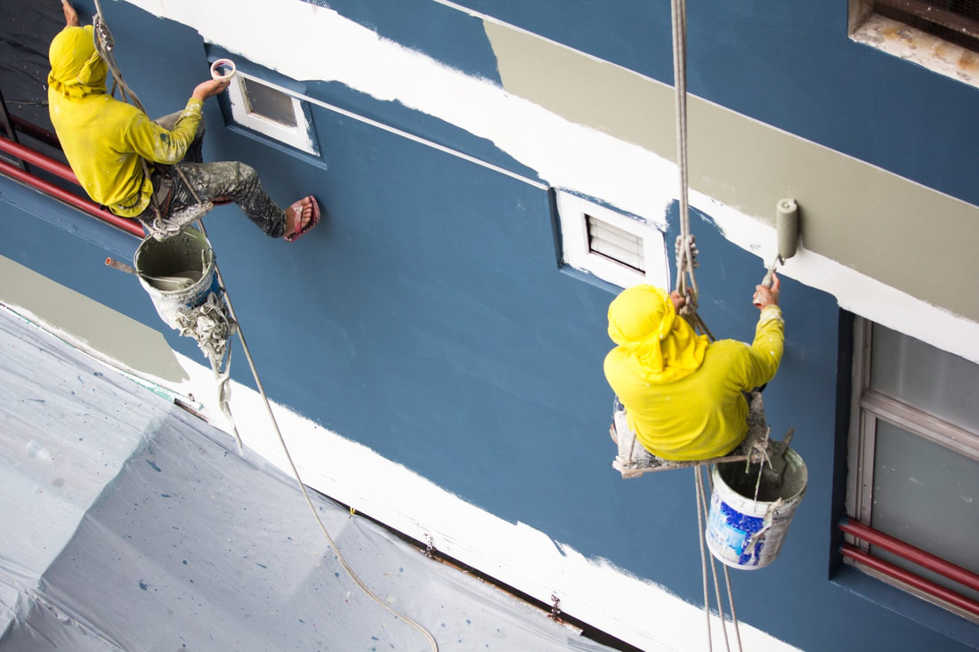 A picture of two painters on ropes painting a huge blue house with gray paint.